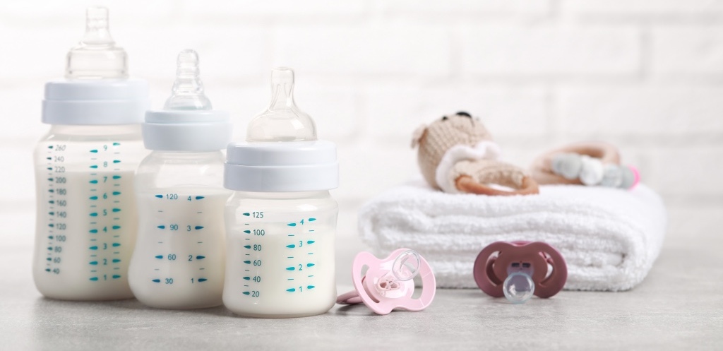 Feeding bottles with milk, pacifiers, toys and towel on light grey table